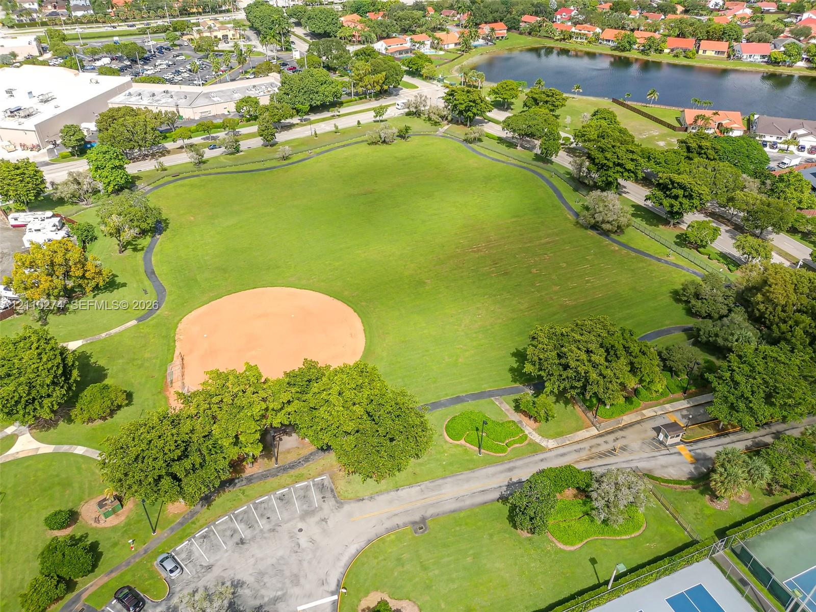 Expansive recreational park grounds featuring a baseball diamond, paved walking paths, paved surface parking, and mature trees