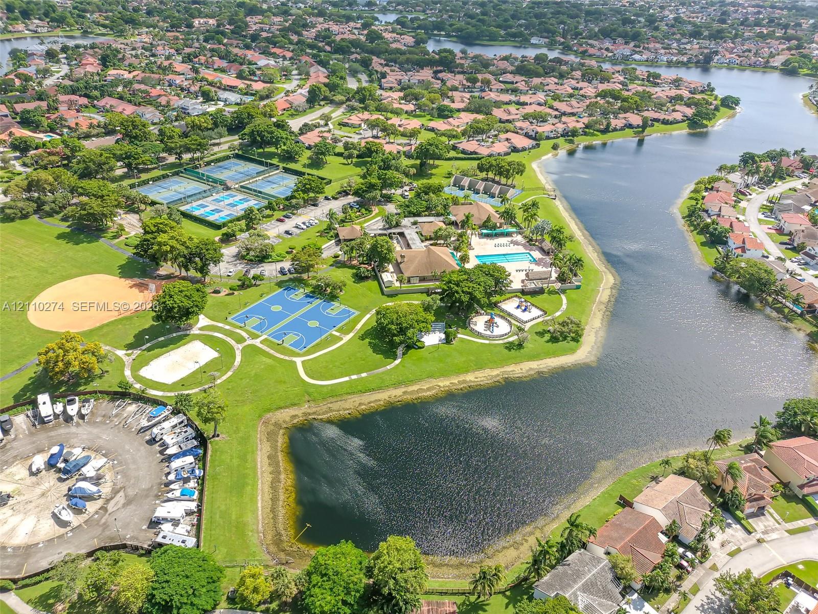 Aerial perspective of community grounds featuring a swimming pool, tennis courts, basketball court, baseball diamond, and boat storage area situated alongside a lake