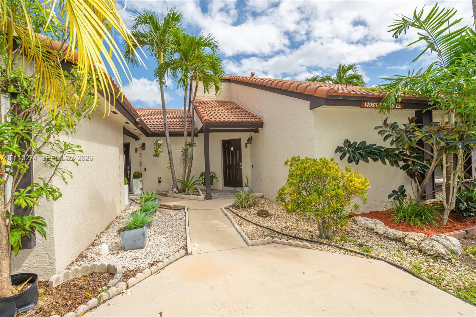 Stucco exterior featuring a clay tile roof, covered entryway, and xeriscaped front grounds with mature palm trees