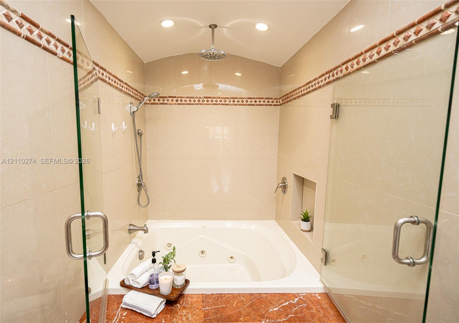 Jetted soaking tub featuring a stone-surround deck, ceiling-mounted rainfall shower head, handheld sprayer, glass enclosure, and decorative tile inlay