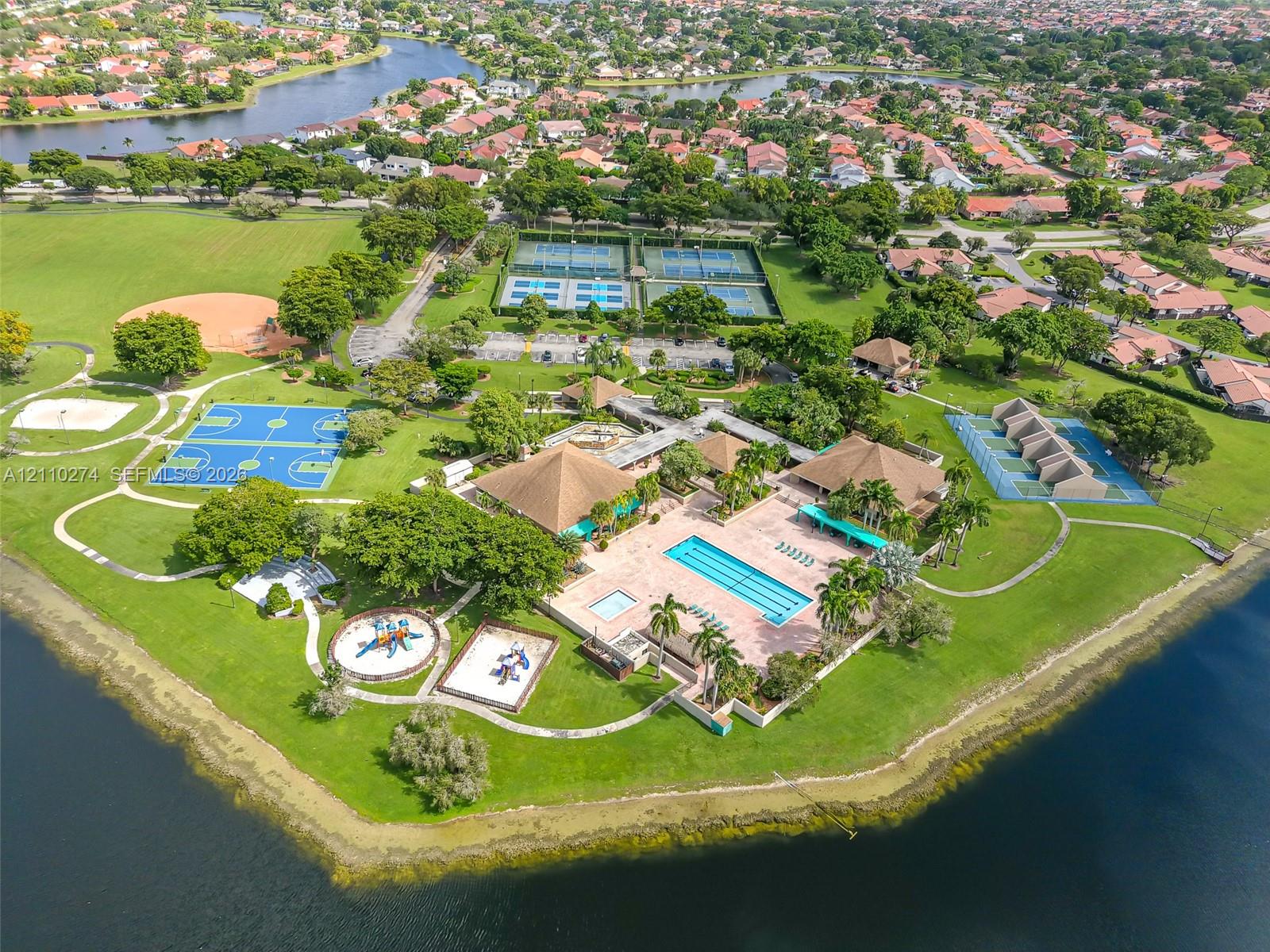 Recreational facility featuring a swimming pool, multiple tennis courts, multi-purpose sports courts, paved walking paths, and mature landscaping situated along a waterfront perimeter