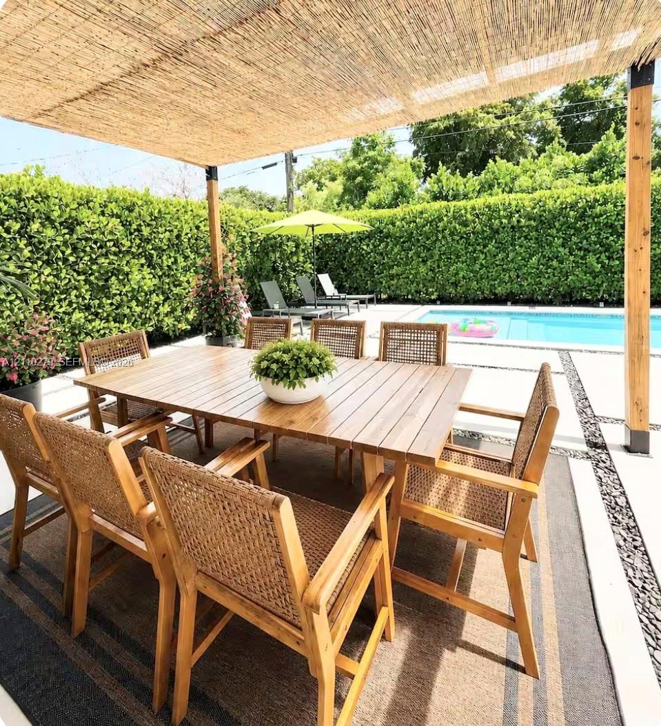 Covered patio featuring a woven reed pergola, concrete paver decking, stone aggregate inlay, and a rectangular swimming pool surrounded by a manicured hedge