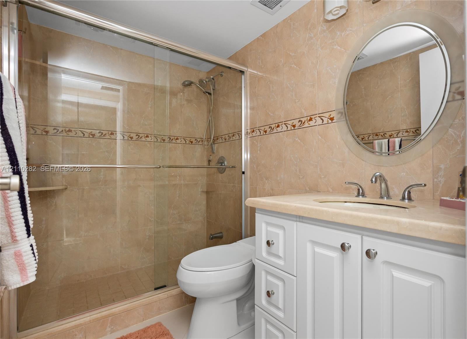 Bathroom featuring a white vanity with a neutral countertop, an integrated sink, a circular wall-mounted mirror, and a walk-in shower enclosure with tile surround and decorative accent border