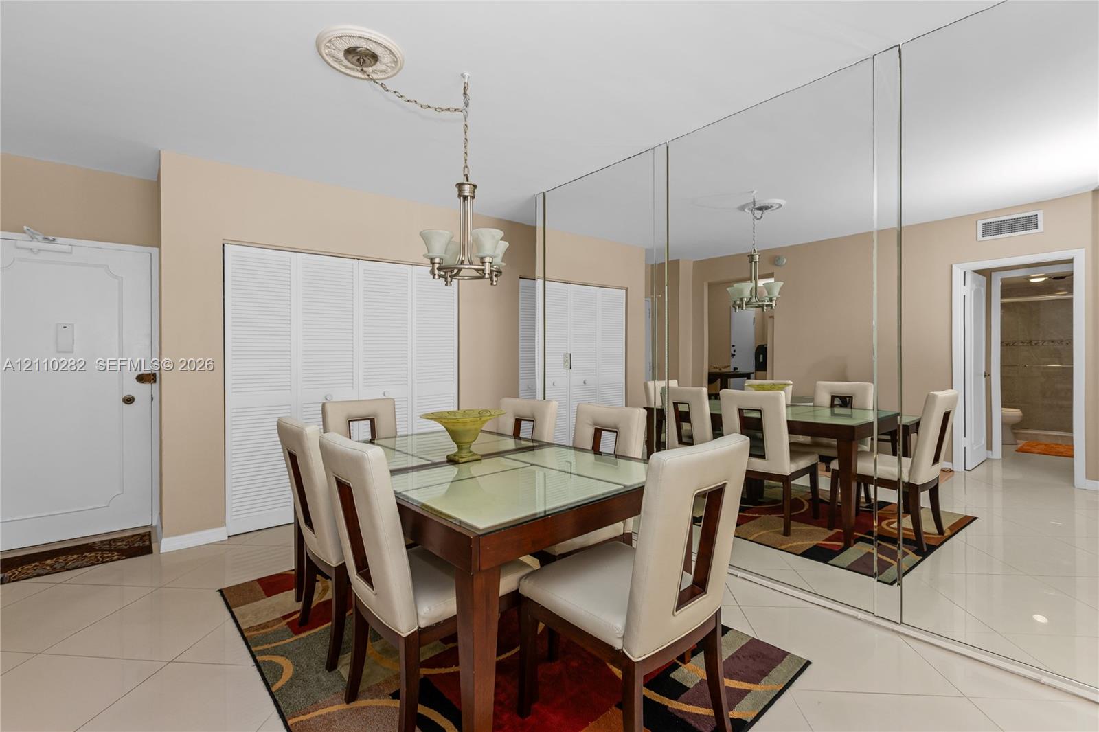 Large-format light-colored tile flooring, full-wall reflective mirror paneling, ceiling-mounted multi-light fixture with decorative medallion, and built-in closet storage with louvered doors