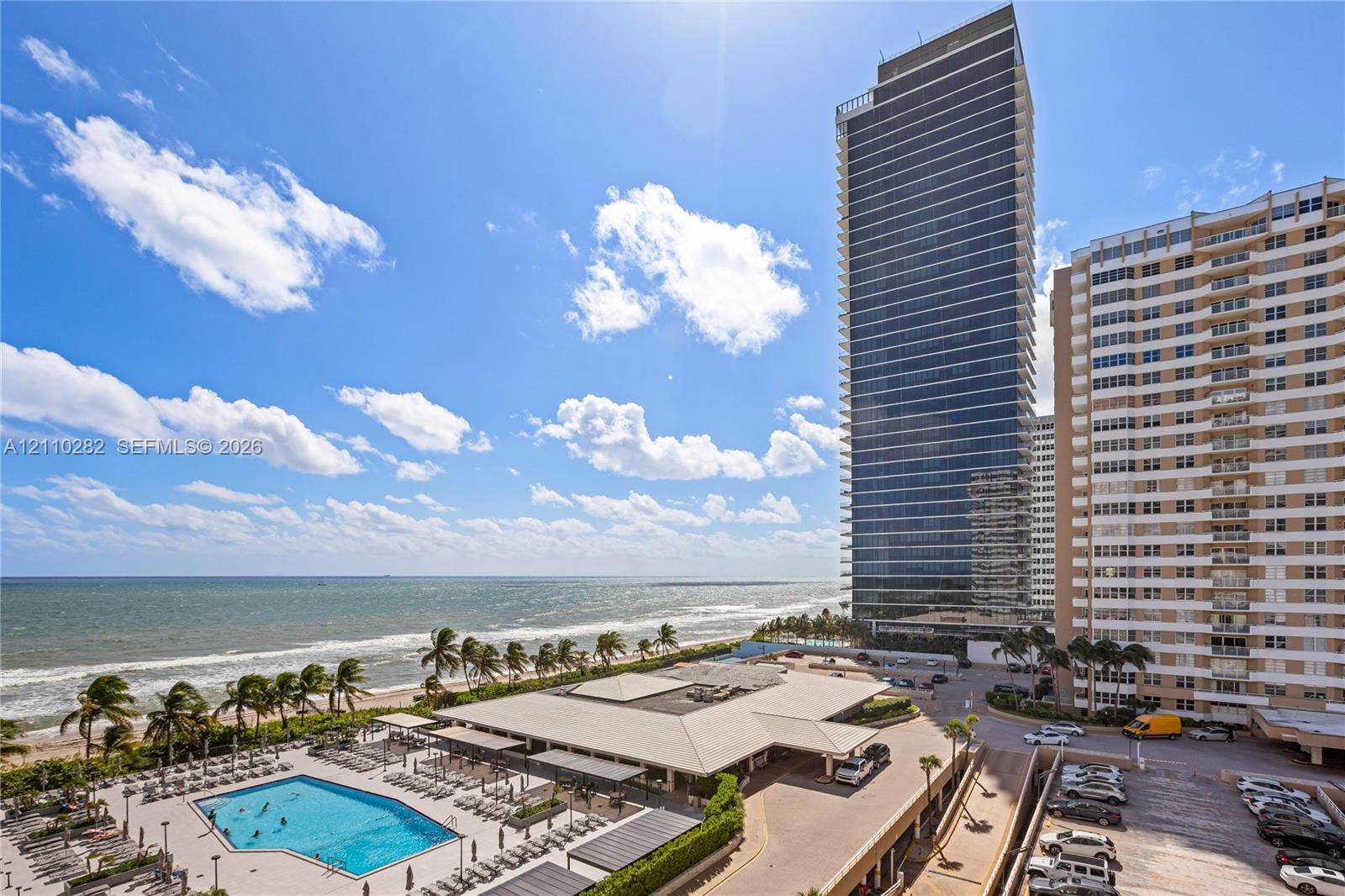 High-rise tower with floor-to-ceiling glass paneling, outdoor swimming pool with paved terrace, and immediate oceanfront location