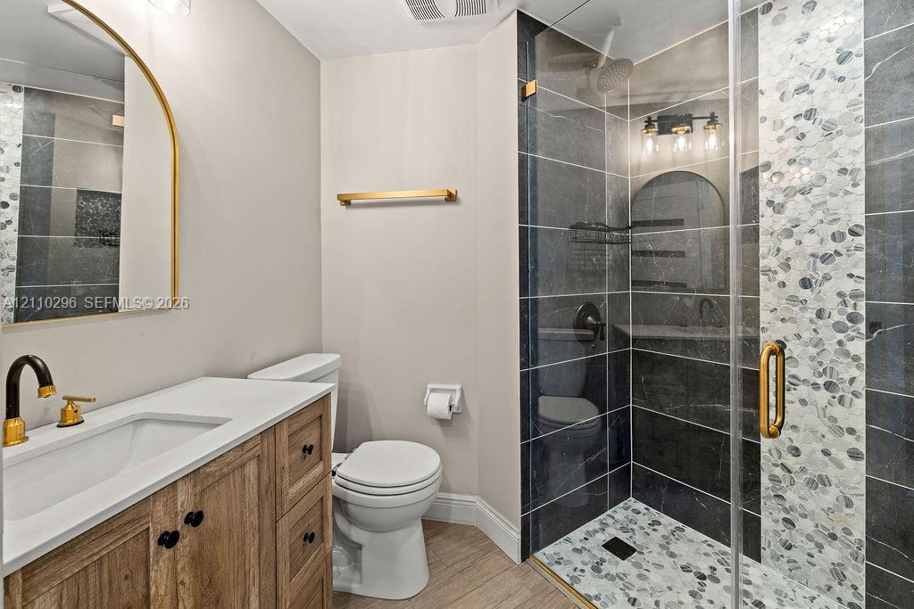 Bathroom featuring a wood-finish vanity with a stone countertop, an arched gold-rimmed mirror, a walk-in shower with stone tile accents, and gold hardware finishes