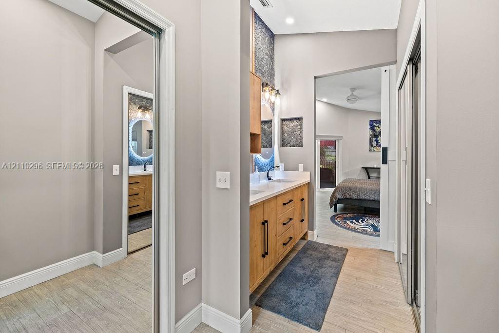Double vanity with wood-finish cabinetry, solid surface countertop, matte black hardware, dual-lit circular mirrors, and textured stone-look tile accent wall