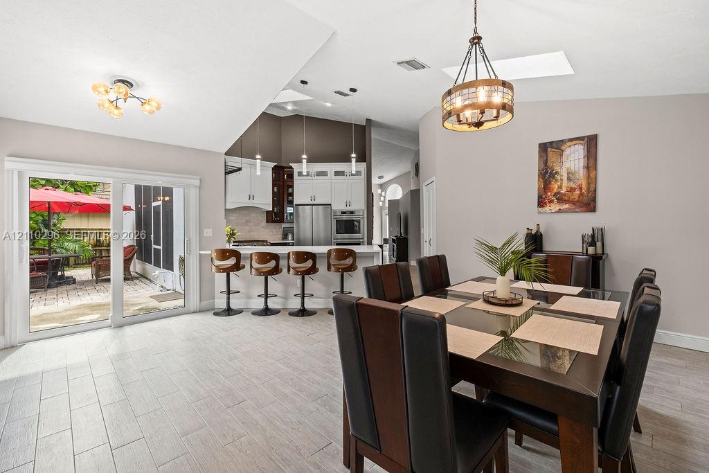 Open-concept interior featuring light-toned tile flooring, vaulted ceilings with skylights, kitchen with white cabinetry and stainless steel built-in appliances, decorative metal and glass pendant lighting, and oversized sliding glass doors