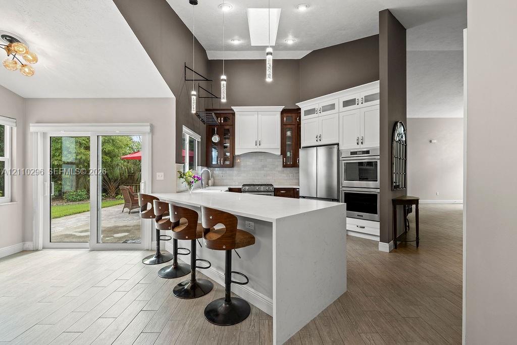 Kitchen featuring an expansive waterfall-edge quartz island, stainless steel double wall ovens, glass-paneled upper cabinetry, tile flooring, and a glass sliding door