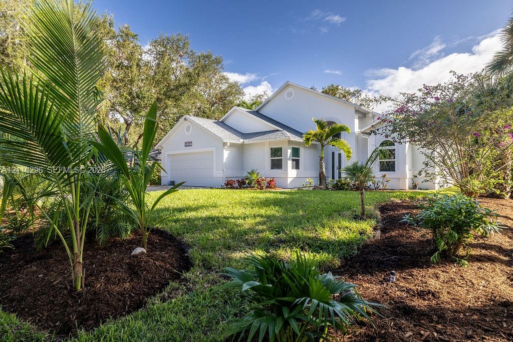 Single-story residence featuring a light-colored stucco exterior, multi-gabled roofline, two-car garage, arched entryway, and mature tropical landscaping