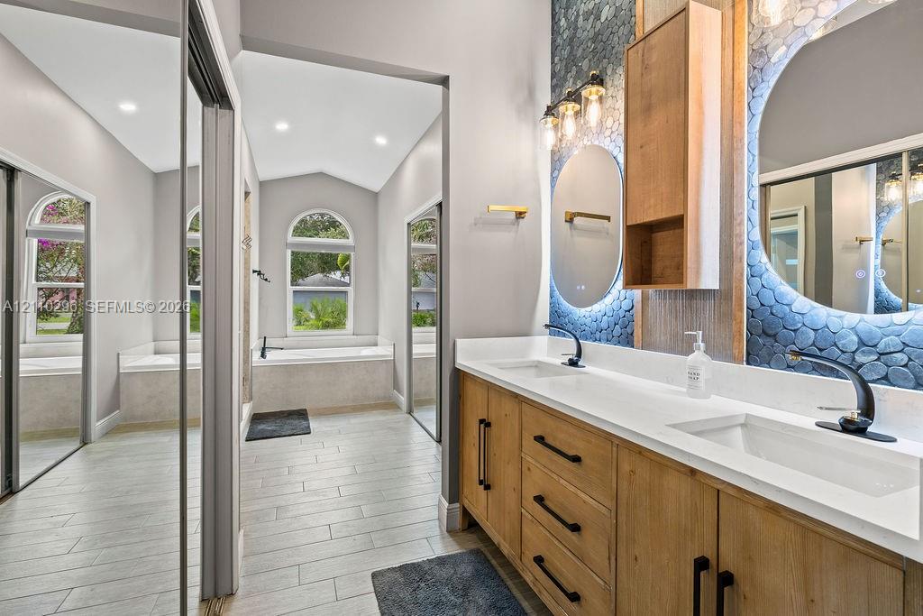 Dual-vanity cabinetry with wood-finish base, quartz countertops, integrated lighting, stone-textured accent wall, and soaking tub with arched window