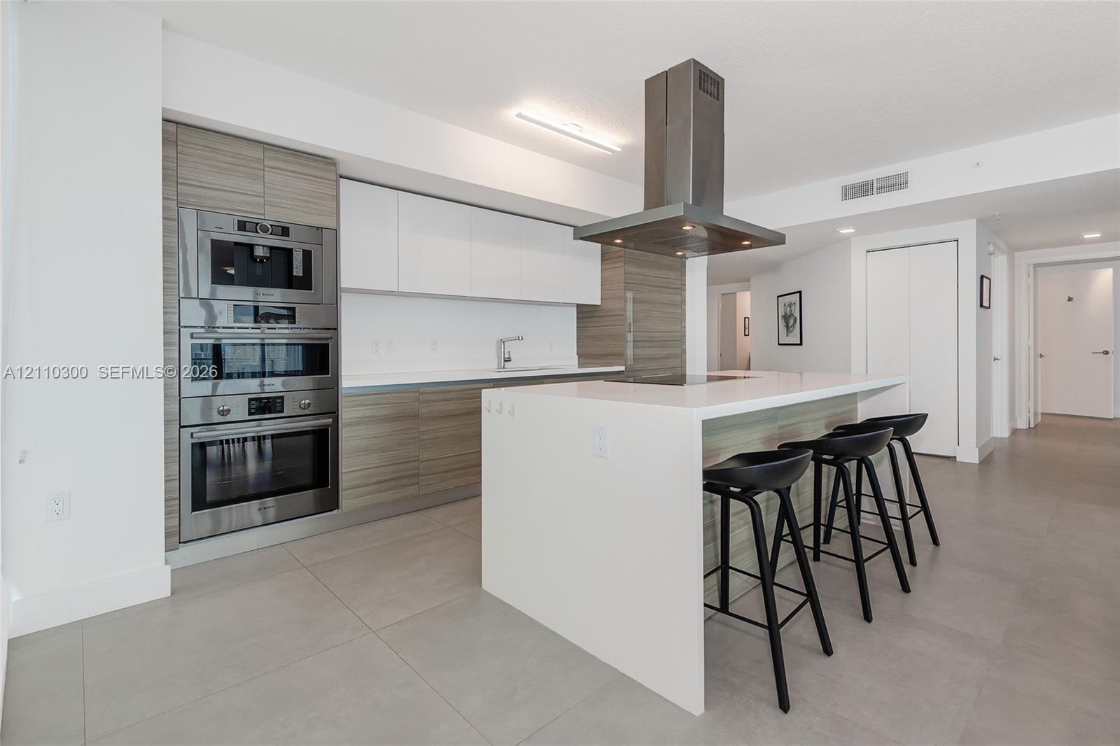 Contemporary kitchen featuring flat-panel cabinetry with wood-finish and solid white fronts, integrated stainless steel double wall ovens, a center island with a white waterfall countertop, and a stainless steel chimney-style range hood