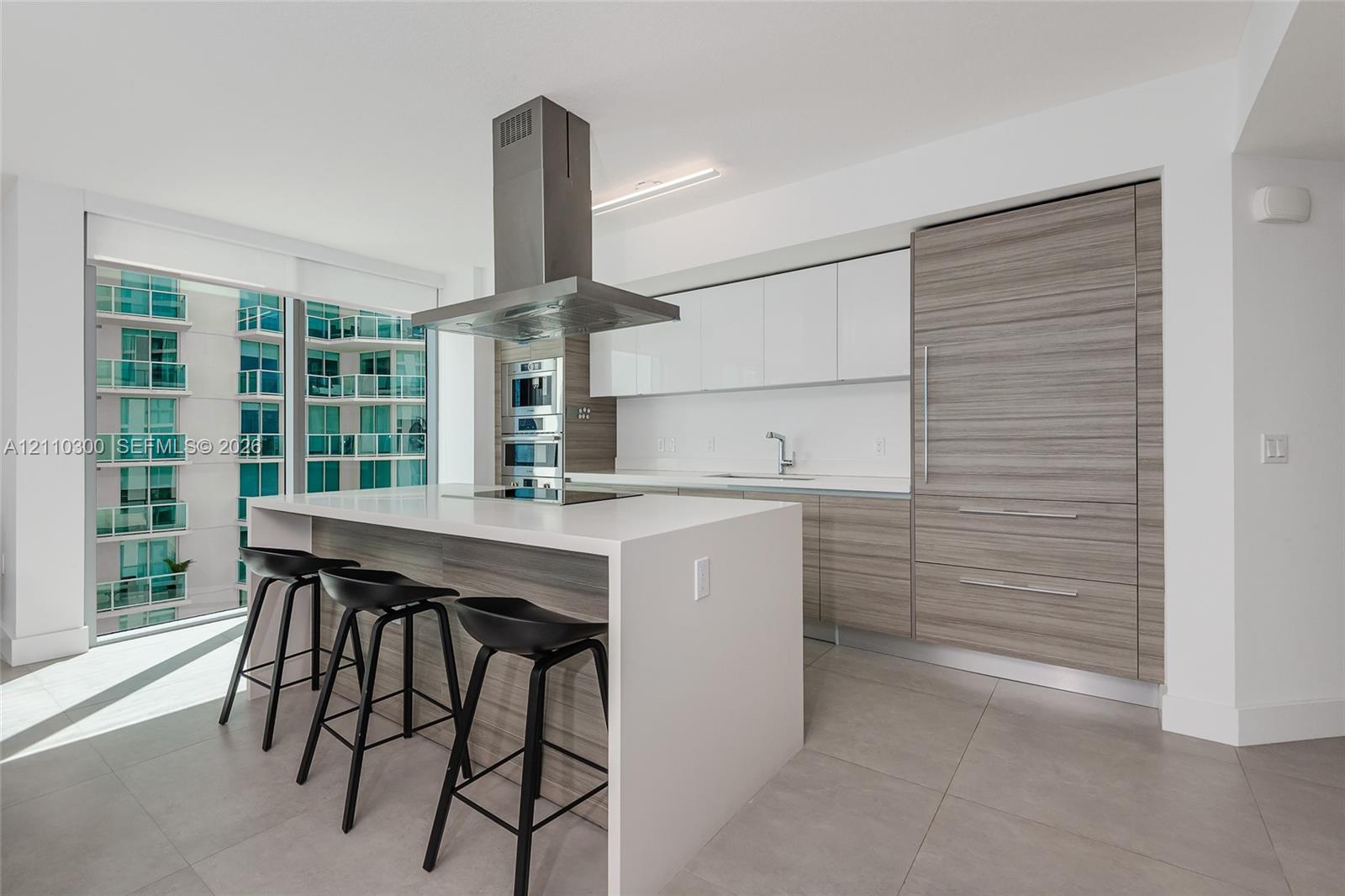 Modern kitchen featuring an island with a waterfall-edge countertop, panel-ready refrigerator, stainless steel ceiling-mounted range hood, built-in double wall ovens, and two-tone flat-panel cabinetry