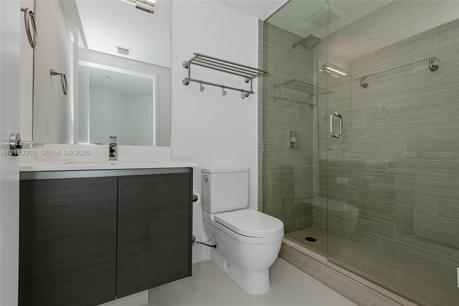 Bathroom featuring a vanity with wood-finish cabinetry and a white countertop, walk-in shower with a glass enclosure and textured tile surround, wall-mounted metal shelving, and a dual-flush toilet