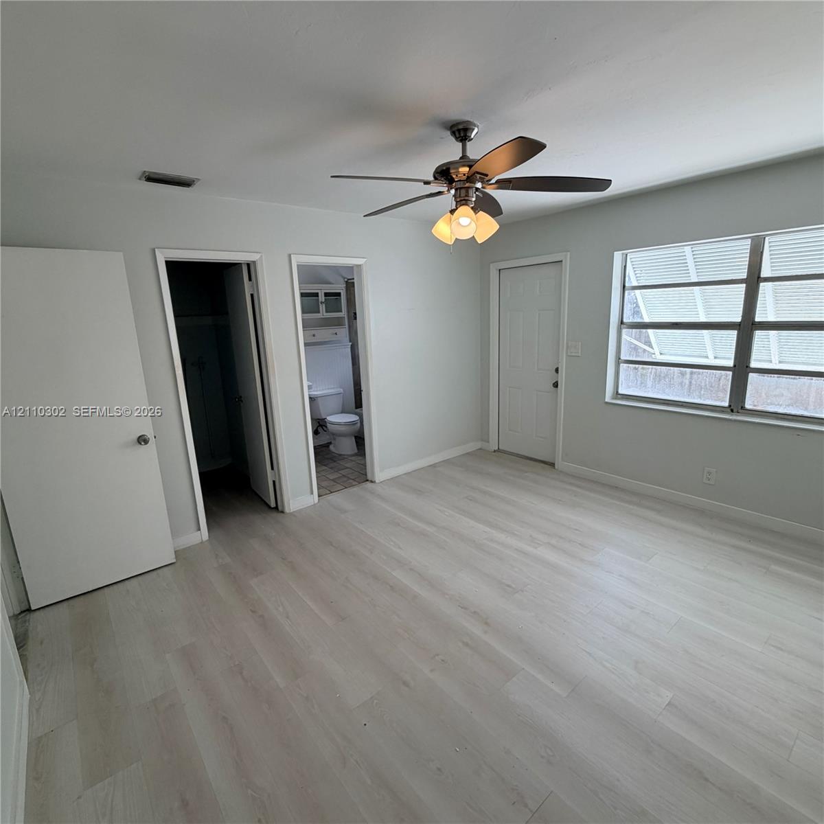 Light-toned wood-finish flooring, central ceiling fan with multi-light fixture, and multi-pane window with adjustable louvers
