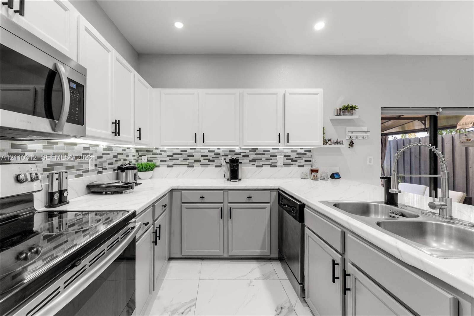 Kitchen featuring white and gray cabinetry, stone-finish countertops, multi-toned glass tile backsplash, stainless steel appliances, and large-format tile flooring