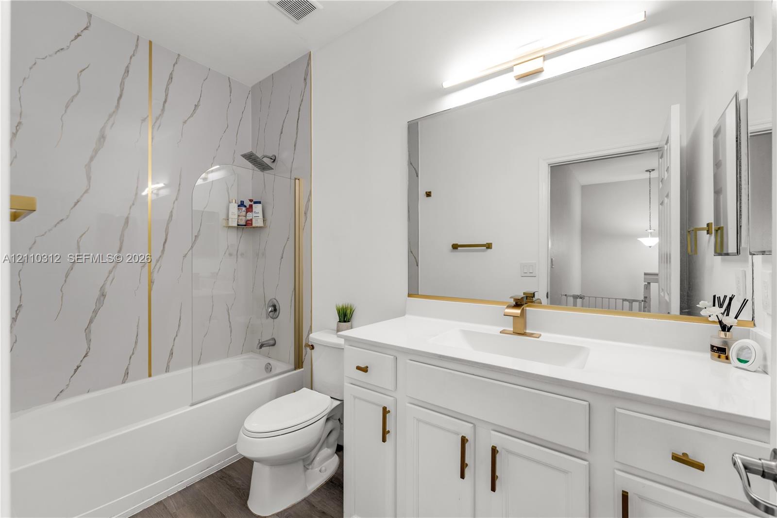 Bathroom featuring a white vanity with gold-finish hardware, a large frameless mirror, a shower-tub combination with gold-accented wall panels, a glass panel screen, and wood-finish flooring