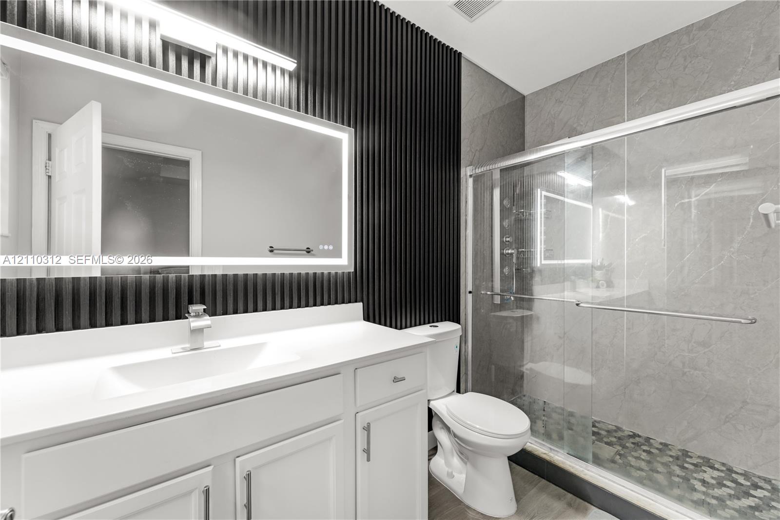 Bathroom featuring a white vanity with a rectangular sink, wall-mounted backlit mirror, black vertical slat accent wall, and a walk-in shower with grey tile surround and stone-style flooring