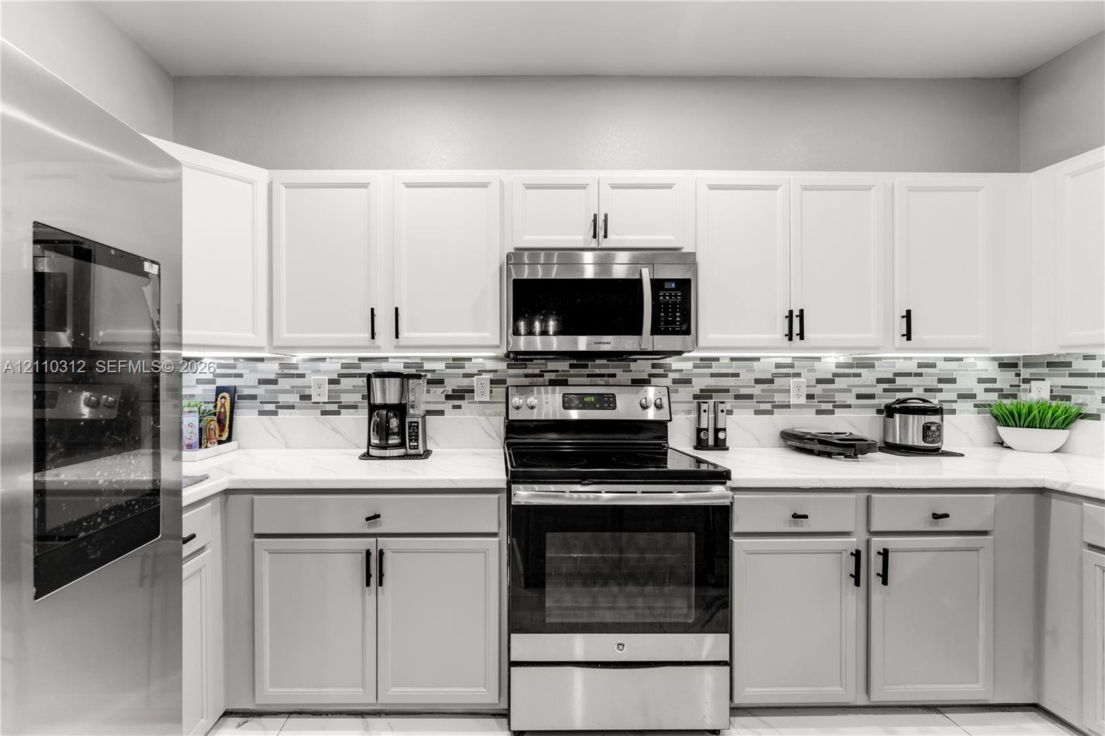 Kitchen featuring white shaker-style cabinetry, solid-surface countertops, a multi-tone glass tile backsplash, and stainless steel appliances
