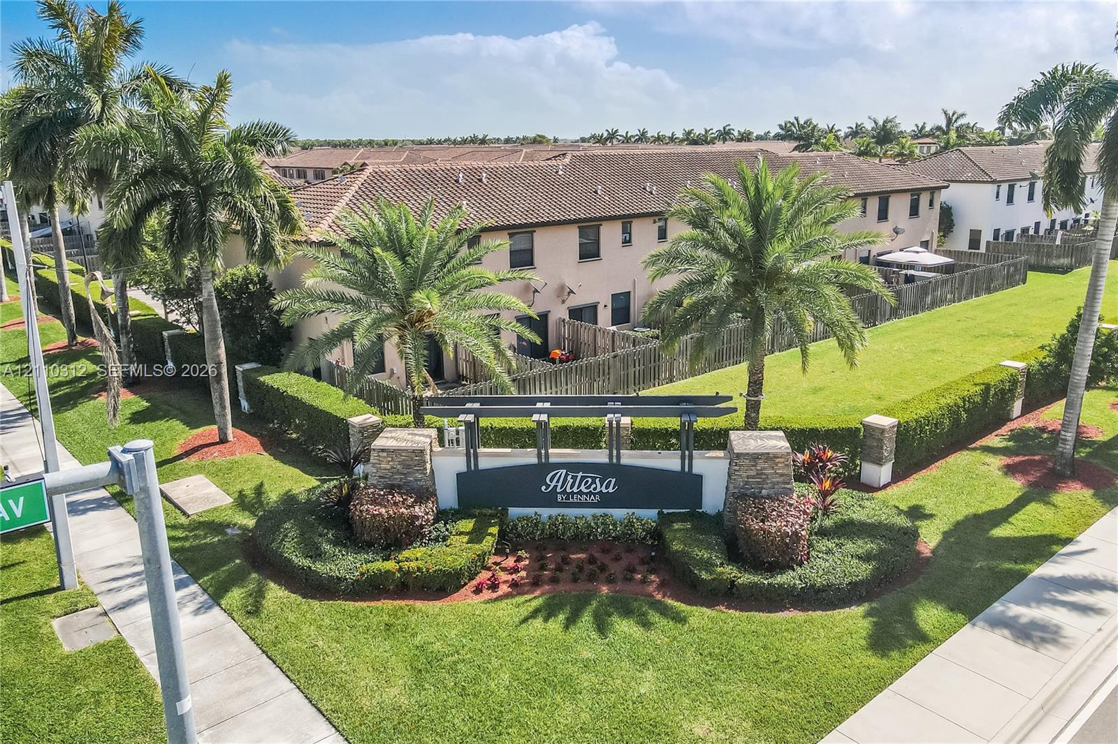 Community entrance sign featuring stone-veneer pillars and metal framing, surrounded by manicured landscaping, mature palm trees, and expansive green space