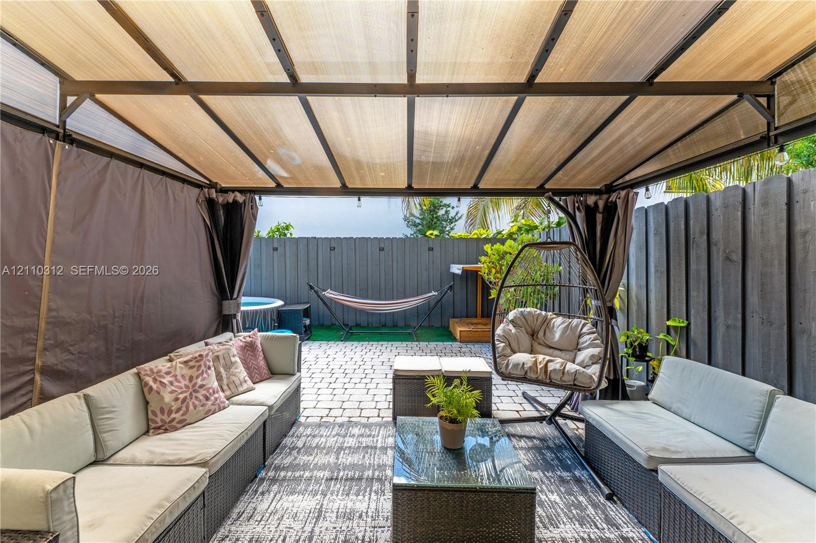Outdoor living space featuring a metal-frame pavilion with a semi-transparent roof, paver patio, and full-perimeter vertical wood fencing