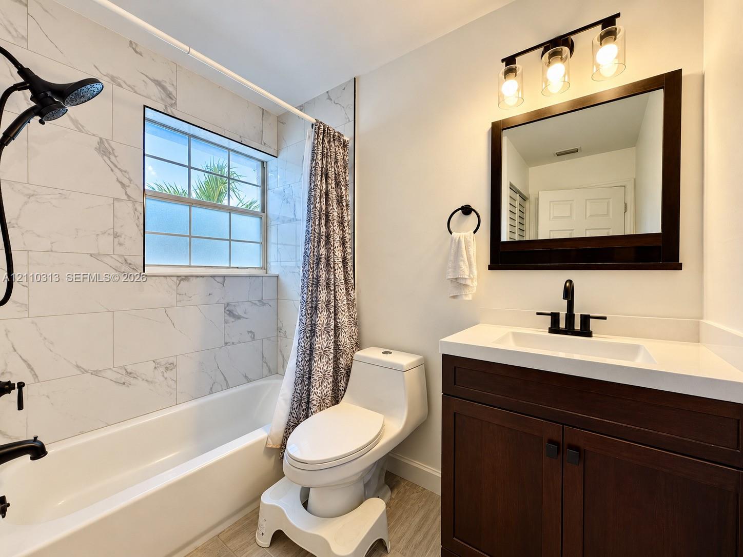 Bathroom featuring a vanity with a white countertop, matte black faucet, wall-mounted mirror, three-light fixture with glass shades, and tub surround tiled in a stone-pattern finish