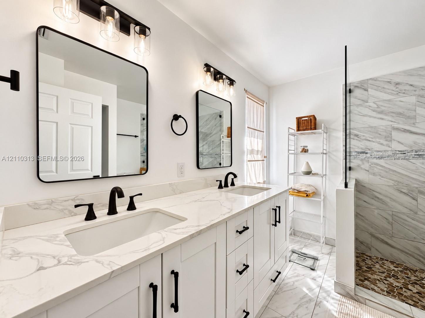 Double vanity featuring white cabinetry, stone-surface countertops, under-mount rectangular sinks, and matte black hardware, accompanied by a glass-enclosed walk-in shower with tiled walls and a stone-tiled floor