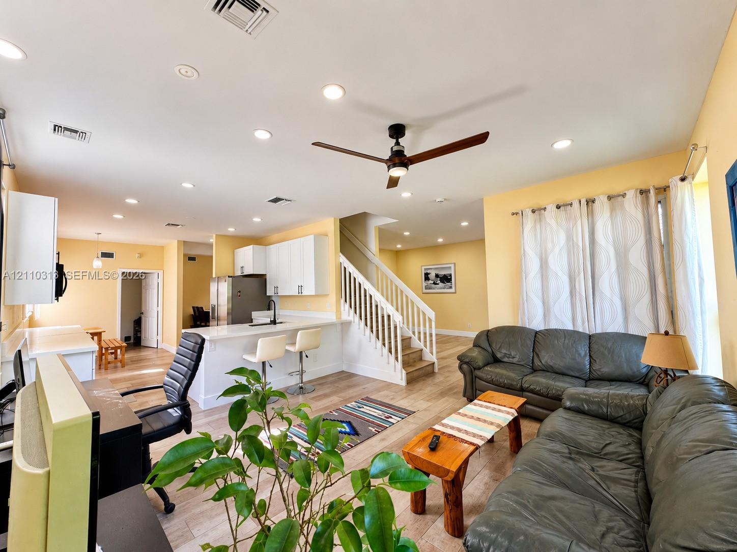 Open-concept interior featuring wood-finish flooring, recessed lighting, a modern kitchen with white cabinetry, a breakfast bar, and a staircase with a white railing