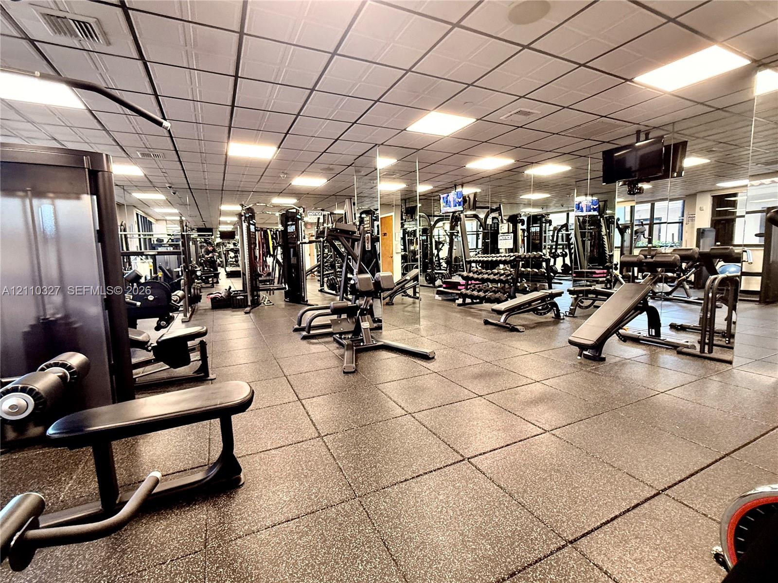 Fitness center featuring grid-pattern acoustic ceiling tiles, recessed lighting, wall-to-wall mirrored surfaces, and high-impact rubber flooring