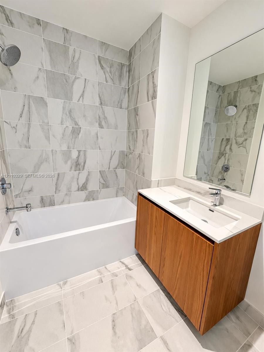 Bathroom featuring a wood-finish vanity with an integrated rectangular sink, stone-composite countertop, full-width wall mirror, and porcelain tile flooring extending to the shower surround