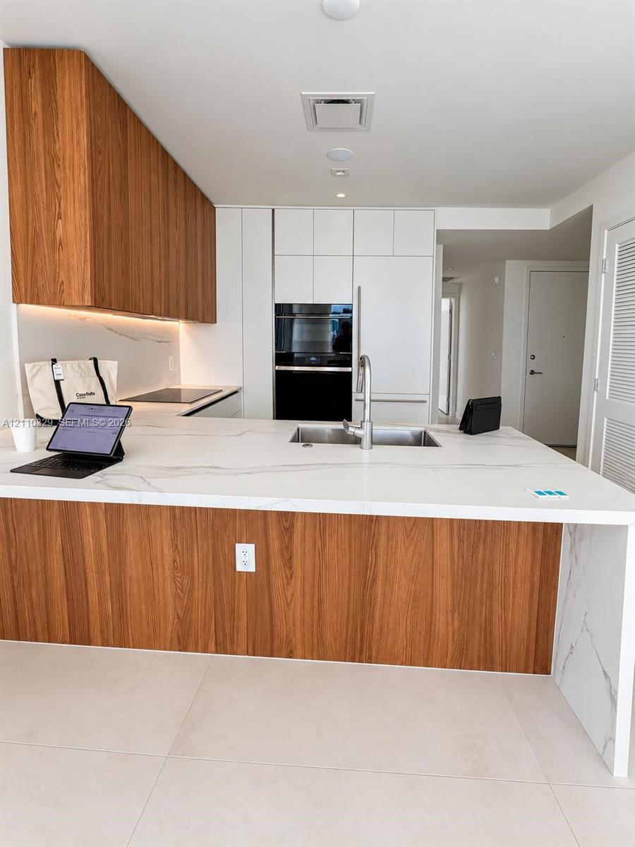 Modern kitchen featuring a waterfall-edge stone countertop, wood-finish cabinetry, built-in wall oven, undermount sink with a gooseneck faucet, and large-format tile flooring