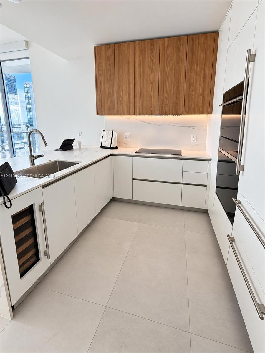 Modern kitchen featuring flat-panel cabinetry with wood-finish uppers, large-format tile flooring, stone-composite countertops, under-cabinet lighting, and built-in stainless steel appliances