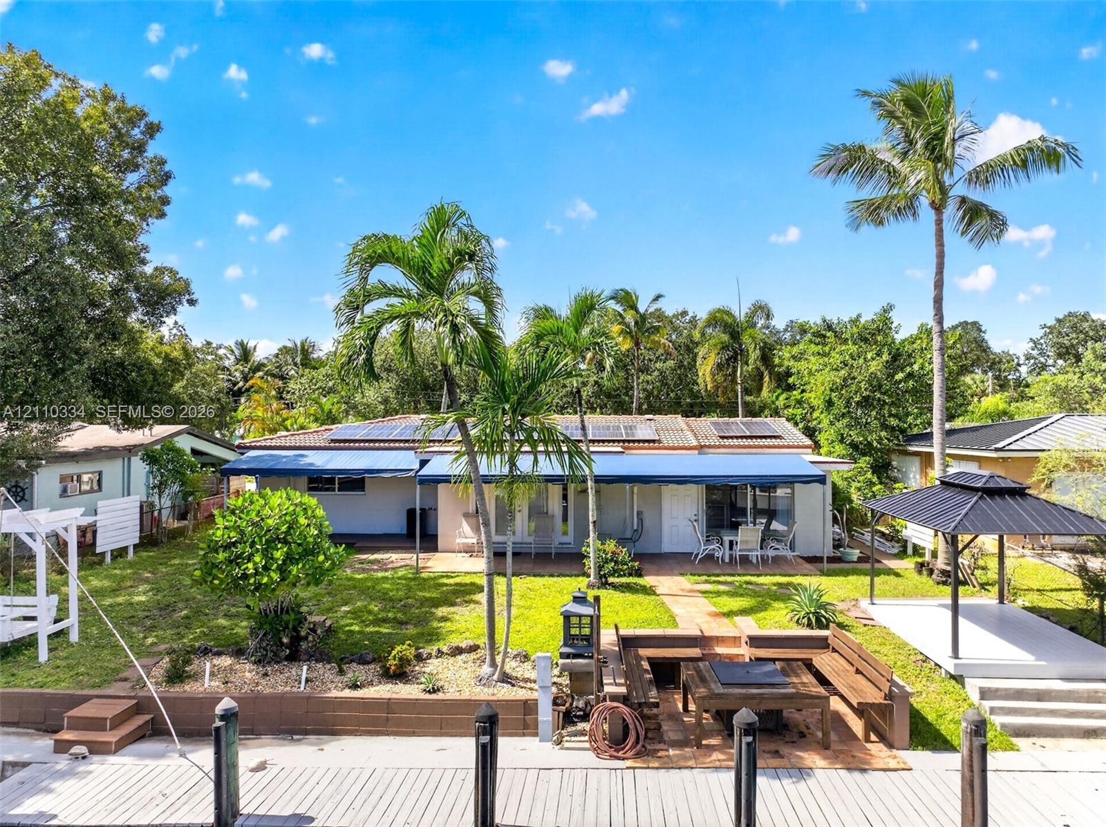 Waterfront backyard featuring a tiered wooden bulkhead, integrated fire pit seating area, covered patio with retractable awnings, detached gazebo, and roof-mounted solar panels