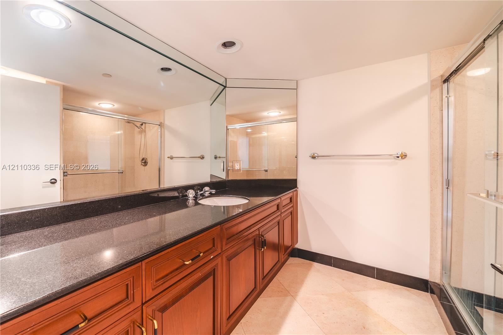 Full-width vanity featuring dark stone-finish countertop, undermount sink, recessed lighting, expansive wall-to-wall mirror, and wood-finish cabinetry