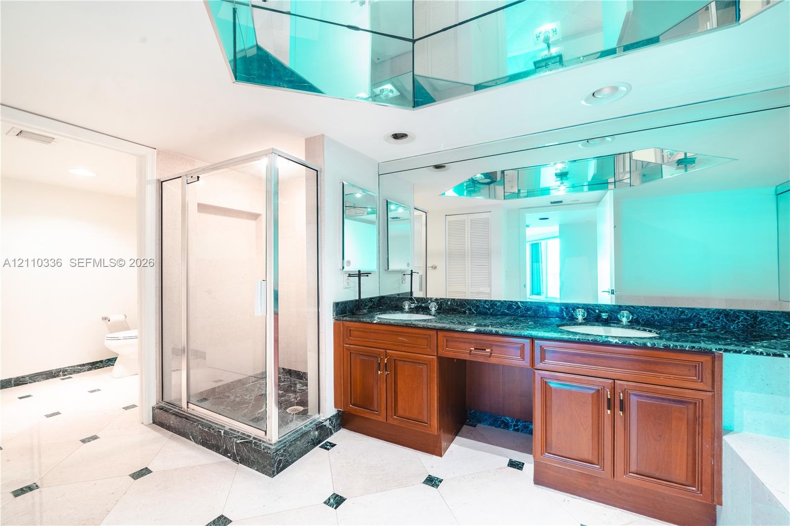 Double vanity with stained wood cabinetry and stone countertops, glass-enclosed shower, expansive wall-to-wall mirror with faceted mirrored accents, and tiled flooring with decorative stone inlays