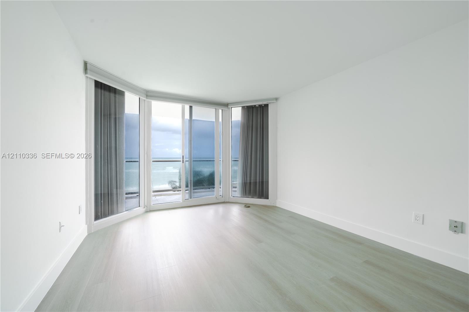 Floor-to-ceiling windows, sliding glass door, wood-finish flooring, and balcony access with water backdrop
