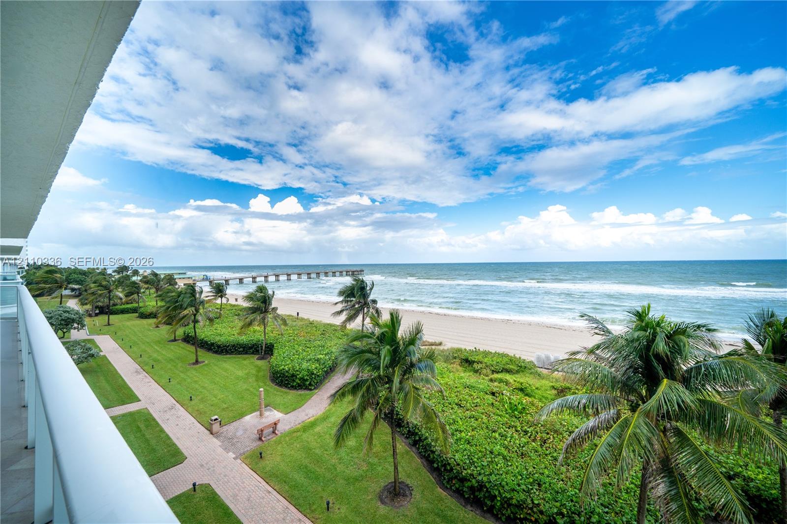 Elevated balcony overlooking coastal frontage featuring palm trees, manicured green space, paved walkways, and a pier extending into the ocean