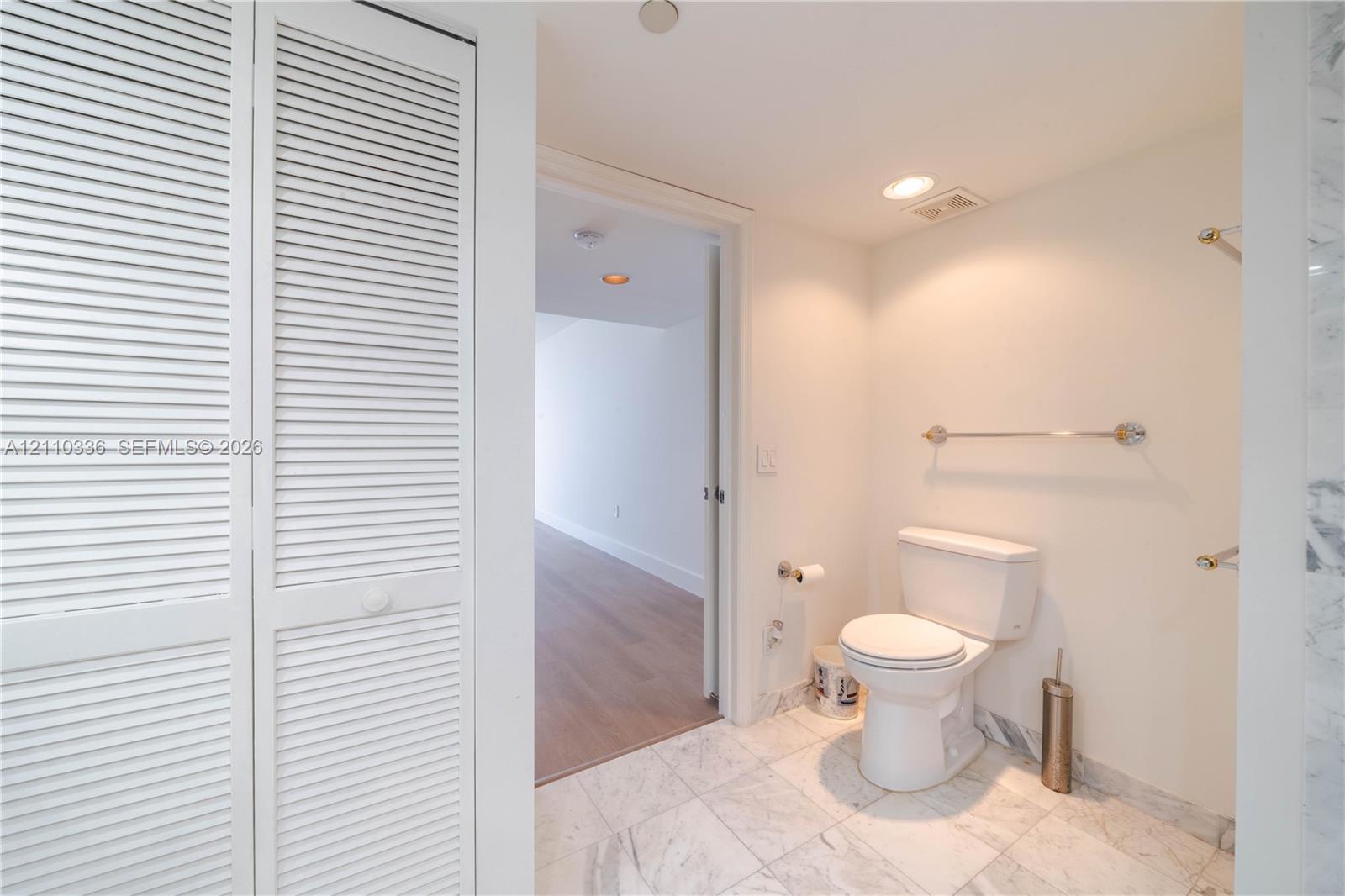 Bathroom featuring white louvered closet doors, stone-patterned tile flooring, a standard-profile toilet, and polished metal wall-mounted towel bars