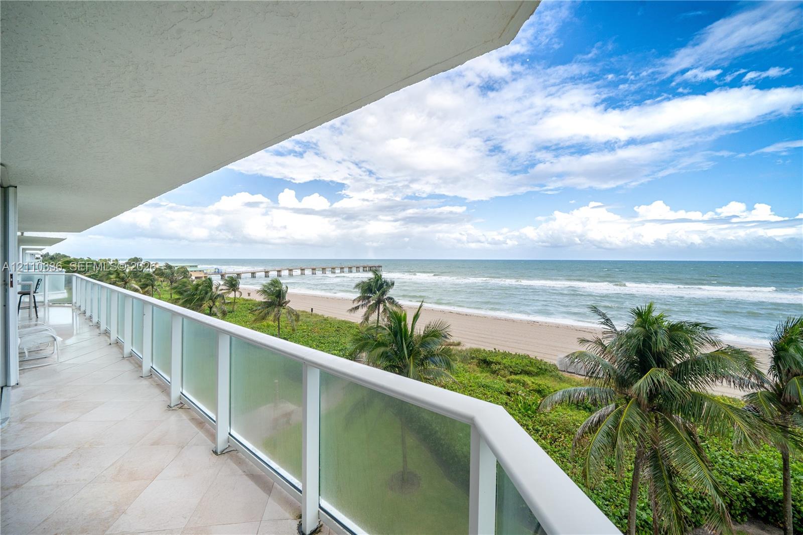 Covered balcony featuring tile flooring and a glass-paneled railing overlooking the ocean, sandy beach, and palm trees Covered balcony featuring tile flooring and a glass-paneled railing overlooking the ocean, sandy beach, and palm trees