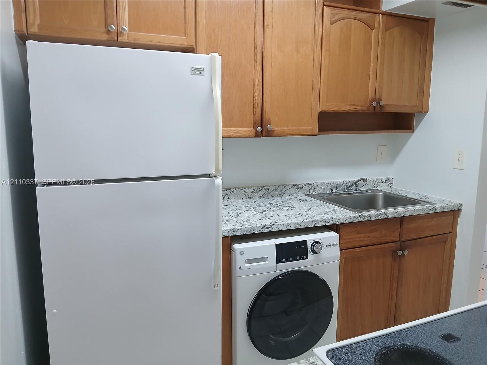 Kitchen area featuring wood-finish cabinetry, stone-patterned laminate countertops, stainless steel sink, and built-in laundry appliance