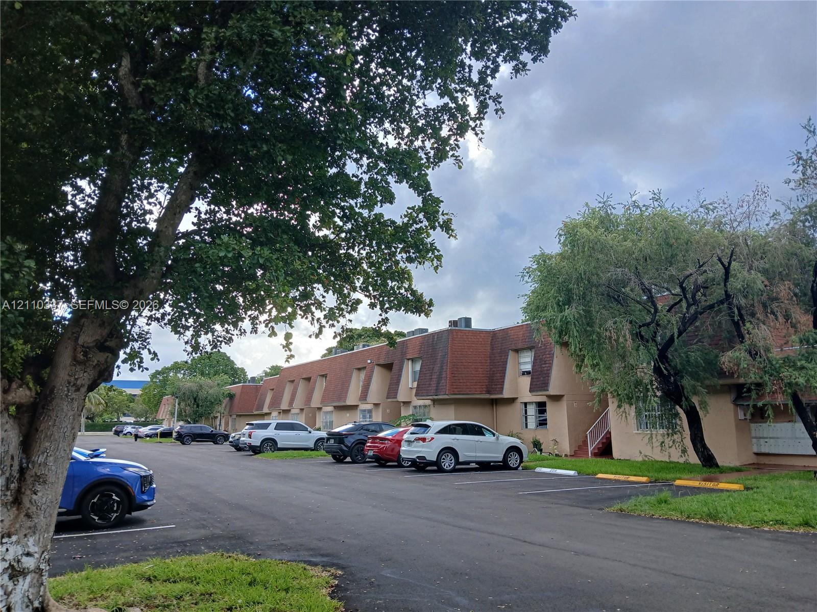 Multi-level exterior featuring a mansard roofline, beige stucco facade, dedicated surface parking, and mature trees Multi-level exterior featuring a mansard roofline, beige stucco facade, dedicated surface parking, and mature trees