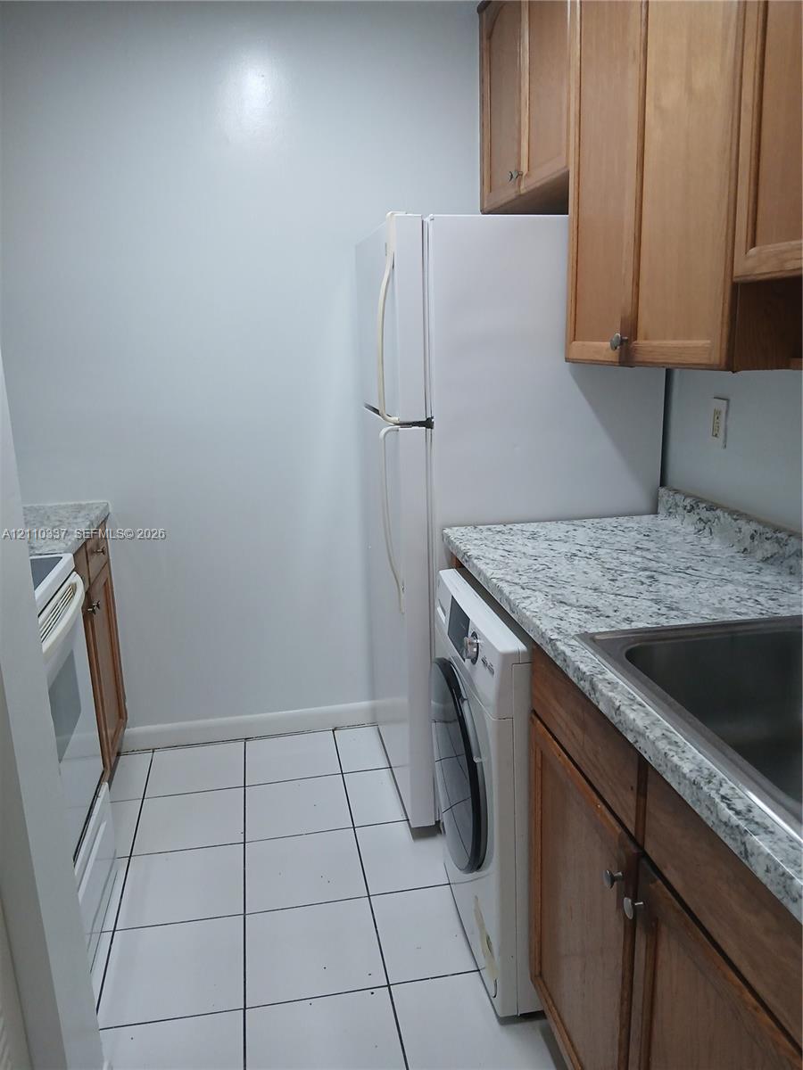 Kitchen featuring shaker-style wood cabinetry, stone-patterned countertops, white ceramic tile flooring, and integrated laundry appliances
