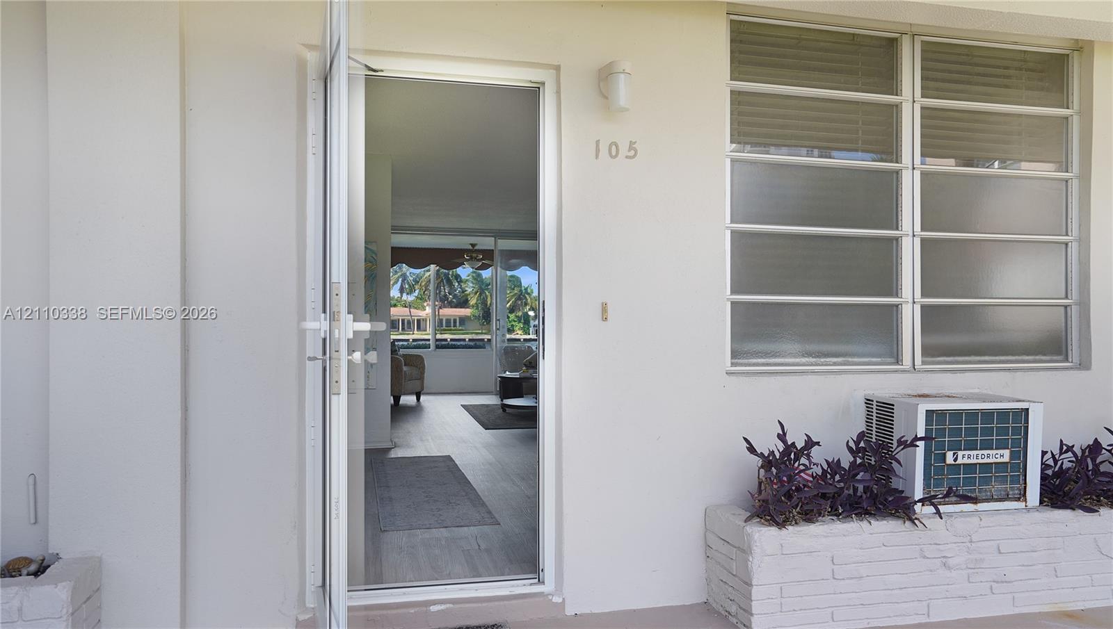 Exterior entrance featuring a white-finish door, louvered glass windows, a mounted exterior light fixture, and a brick-based masonry planter