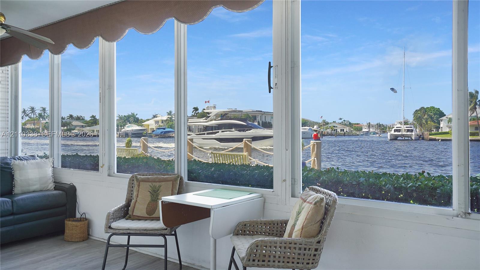 Enclosed sunroom featuring expansive floor-to-ceiling windows, wood-finish flooring, and a retractable fabric awning, overlooking a private canal with bulkhead and nautical access Enclosed sunroom featuring expansive floor-to-ceiling windows, wood-finish flooring, and a retractable fabric awning, overlooking a private canal with bulkhead and nautical access