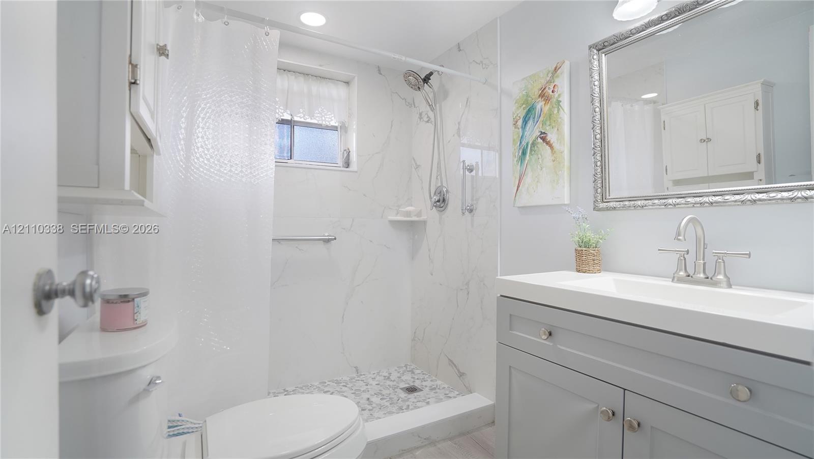 Walk-in shower with stone-finish surround and integrated shelving, grey vanity with rectangular basin, brushed-nickel widespread faucet, and rectangular mirror