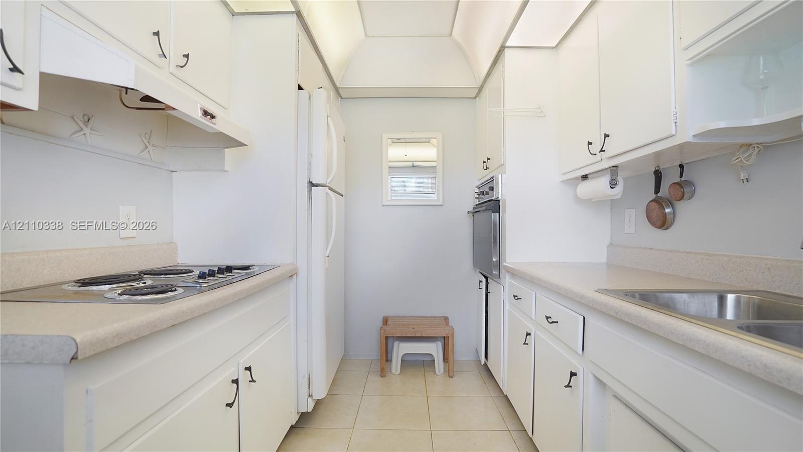 Galley kitchen featuring white shaker-style cabinetry, solid-surface countertops, a stainless steel double-basin sink, an integrated wall oven, and a cooktop