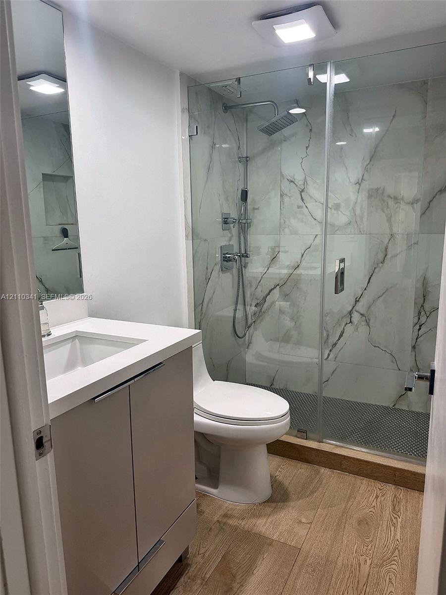 Bathroom featuring a vanity with a white countertop and rectangular sink, frameless glass-enclosed shower with large-format stone-patterned wall tile, rain-style shower head, and wood-finish flooring Bathroom featuring a vanity with a white countertop and rectangular sink, frameless glass-enclosed shower with large-format stone-patterned wall tile, rain-style shower head, and wood-finish flooring