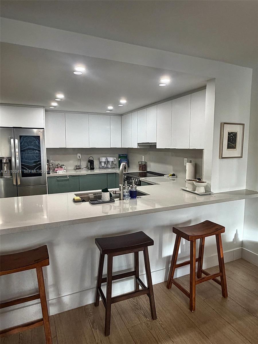 Kitchen featuring two-tone cabinetry, stainless steel refrigerator, stone-surface breakfast bar with integrated sink, recessed lighting, and wood-finish flooring Kitchen featuring two-tone cabinetry, stainless steel refrigerator, stone-surface breakfast bar with integrated sink, recessed lighting, and wood-finish flooring