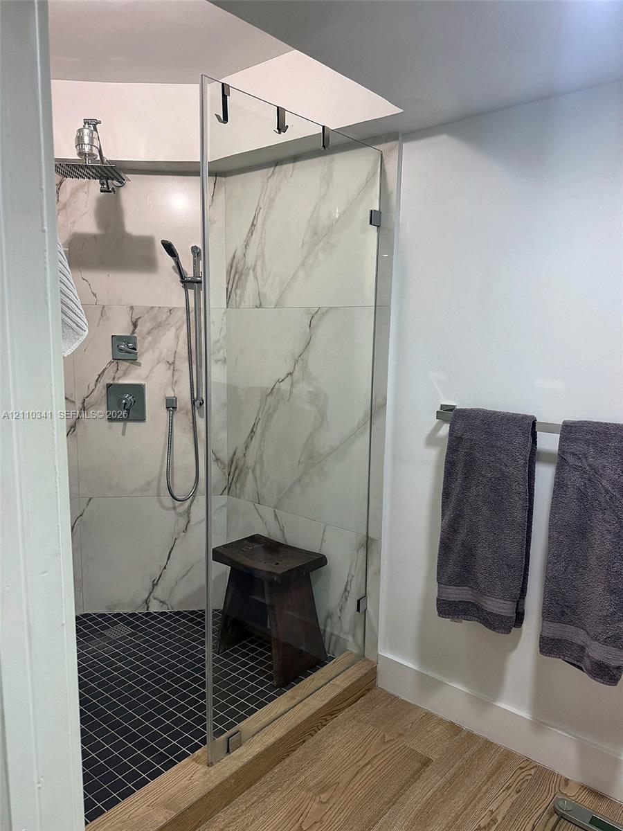 Walk-in shower featuring large-format stone-pattern tile walls, a grid-patterned floor, a frameless glass door, a rainfall showerhead with a handheld attachment, and wood-finish flooring Walk-in shower featuring large-format stone-pattern tile walls, a grid-patterned floor, a frameless glass door, a rainfall showerhead with a handheld attachment, and wood-finish flooring