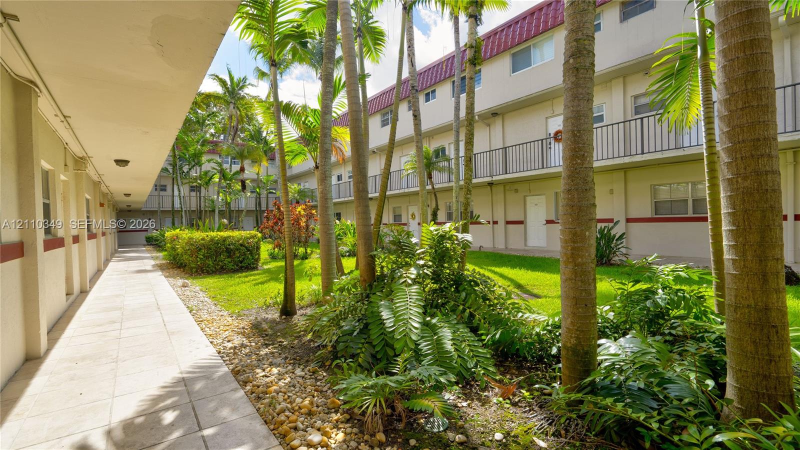 Covered exterior walkway featuring a tiled floor, manicured lawn, stone ground cover, mature palm trees, and multi-level railings